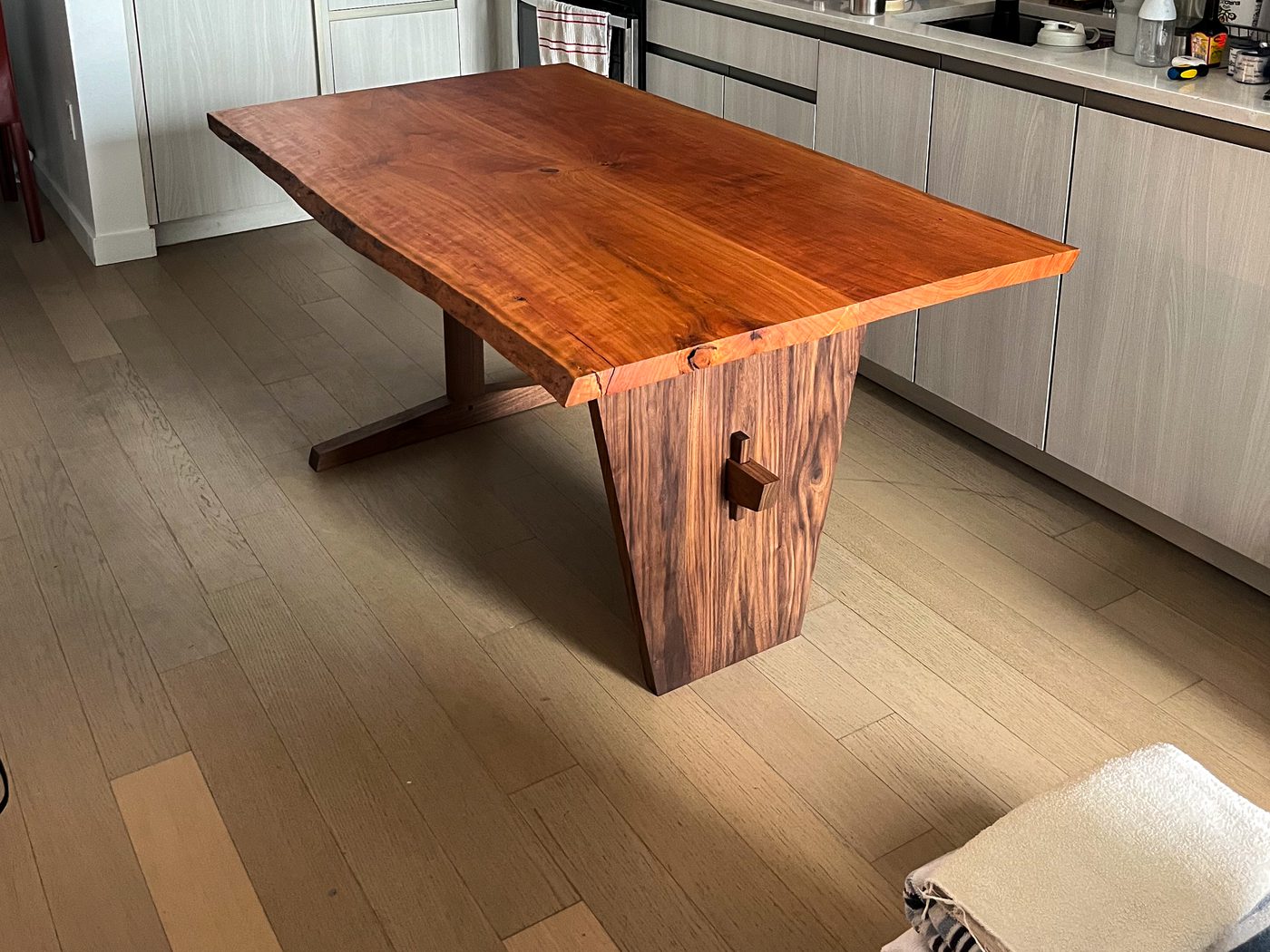 A live-edge walnut dining table with a solid panel leg at one end and an open trestle at the other, a wedged tenon showing through the panel